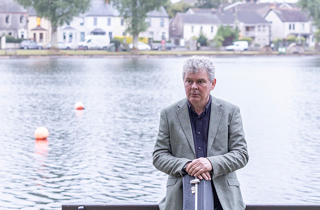 John Spillane stands in front of a river in the daytime, he is wearing a suit jacket and has his hands resting on his guitar case held in front of him.