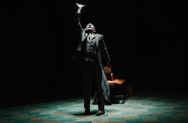 Man in a dark suit stands alone under a spotlight on a stage, reaching one hand upward dramatically beside an orange chair in a dark theatre.
