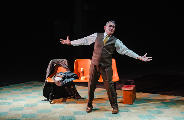 Man on a theatre stage stands with arms outstretched beside orange seats, a draped coat and small wooden box, performing under stage lights.