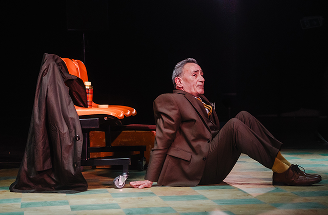 Man in a brown suit sits on a stage floor beside an orange chair with a coat draped over it, gazing ahead under theatrical lighting.