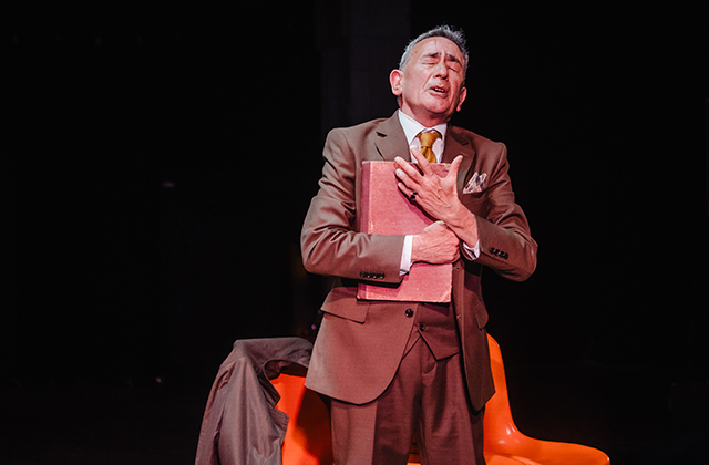 Man in a brown suit stands on stage clutching a red book to his chest, eyes closed in an emotional moment, with an orange chair behind him.