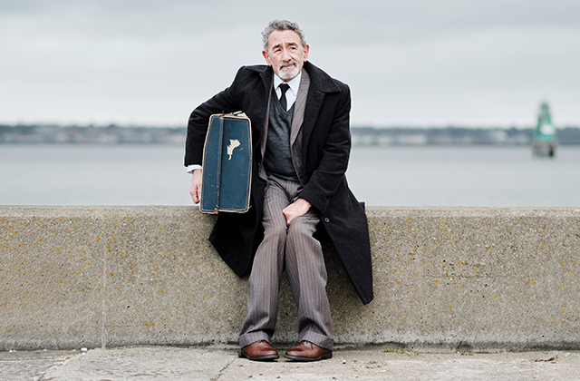 An older man in a dark overcoat sits on a concrete seawall, holding a worn suitcase, with a calm body of water and cloudy sky behind him.