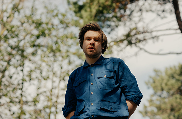 Ronan Kealy AKA Junior Brother stands towards the camera, he is standing outside in the daytime, surrounded by trees, he wears a blue shirt, the camera is below him & he looks down towards the camera.