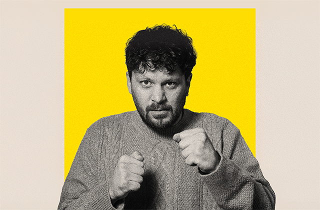 A black-and-white portrait of a Killian Sundermann - a  man with curly dark hair and a short beard - wearing a thick knit jumper, holding his fists up in a playful boxing stance against a bright yellow background.
