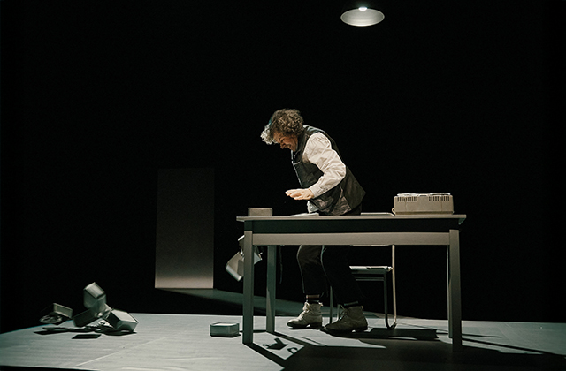 Stephen Rea, as Krapp, appears to have just smashed a machine on the ground in front of him. He has a look of exertion on his face. Behind him is chair, and in front of him is a table.