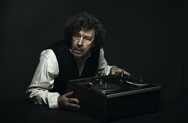 Stephen Rea, as Krapp, listens intently to an old reel-to-reel tape machine. He wears a black waistcoat over a white shirt, and the background is pitch black.