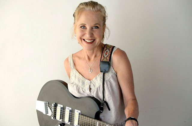 Kristin Hersh stands centre shot, she is against a grey background, she is standing toward the camera smiling, she is holding her guitar which is hanging off her shoulder.