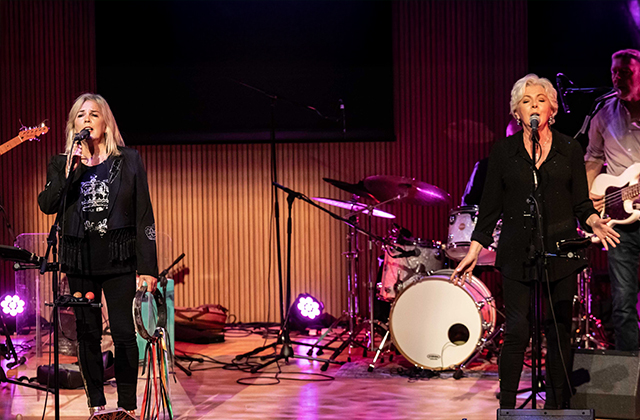Leslie Dowdall and Flo McSweeney stand on opposite sides of a stage, with a full band around them. Both are singing passionately into microphones.