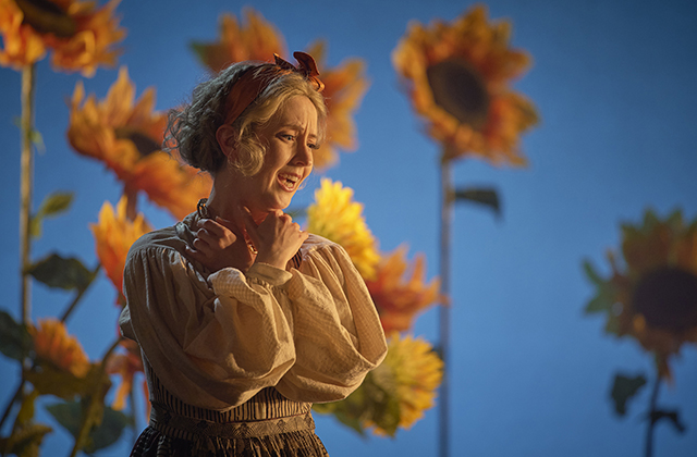 A woman stands staring down with a worried expression on her face. In the background there are sunflowers. Sarah Richmond from Irish National Opera L'Olimpiade Ros Kavanagh