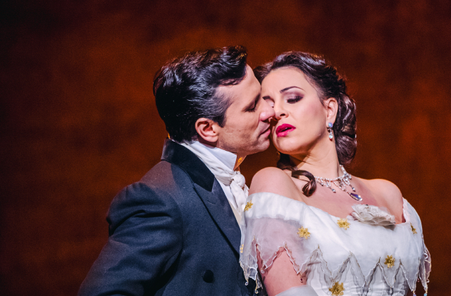 Two opera performers in period costumes share an intimate moment onstage. The man, dressed in a dark formal suit with a white cravat, leans in as if to kiss the woman’s cheek. The woman, wearing an elegant off-the-shoulder white gown with gold accents and ornate jewelry, tilts her head back with a dramatic, emotional expression. Warm, theatrical lighting highlights their faces against a blurred background.