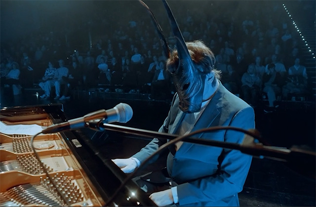 Lambert sits at the piano with his trademark mask he is wearing a suit, the audience is in the background. The light is very blue