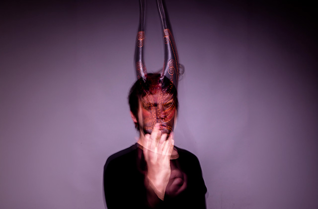 The masked pianist stands in the centre of the image, against a dark, purple and misty background. The large horned mask covers his face somewhat, but is also translucent to see his face at the same time, but neither the face or mask is clear