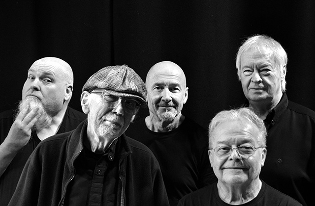 Five members of Lindisfarne are standing looking toward the camera. Rod Clements where's a flat cap with glasses and is smiling. The rest of the band stand around him in a black and white image.