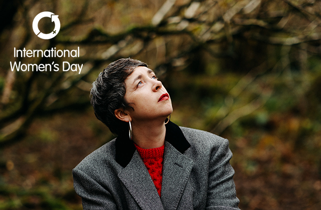 Lisa O'Neill wearing a grey blazer and red jumper, looking wistfully to the sky, with a forest behind her.