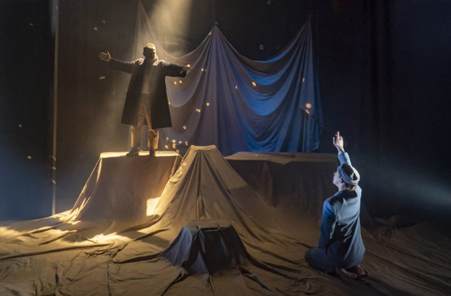 A dramatic scene of a silhouetted man with arms outstretched standing on a table, while in the foreground a young woman points to the ceiling.