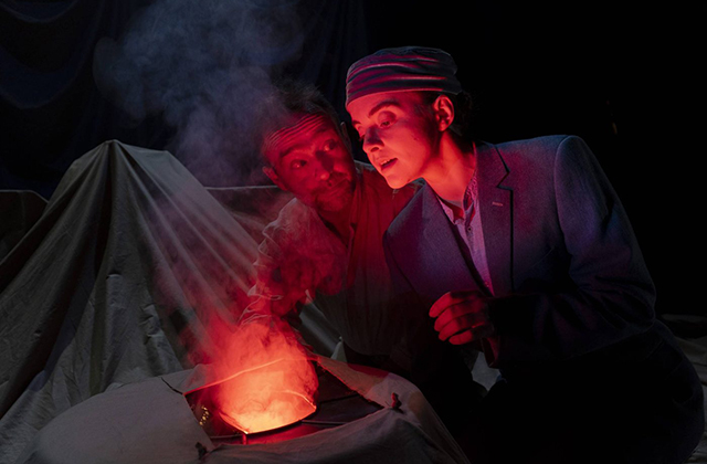 Dramatic shot of two actors an older man and younger women, huddled around what looks like a fire.