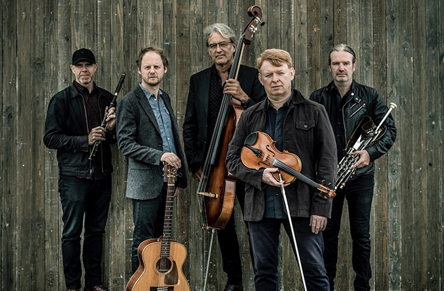 Five men stand against a weathered wooden wall, holding traditional instruments including fiddle, flute, uilleann pipes, guitar and double bass, dressed in dark, casual clothing.