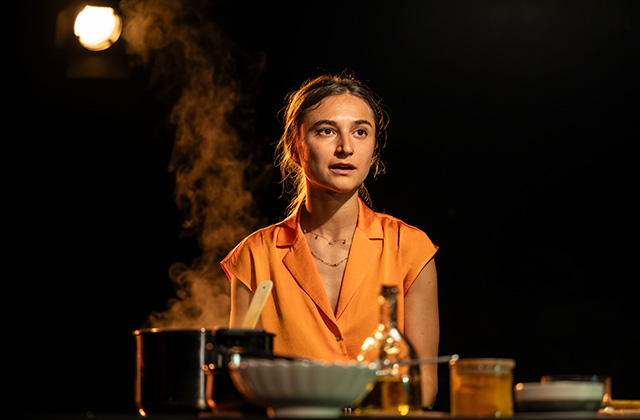 Isabella Nefar, in My English Persian Kitchen. The woman stands in the entre of the image, with a bright orange shirt. In front of her is a counter top with various cooking bits. The background is dark with a bright light emanating from the top left of the picture.