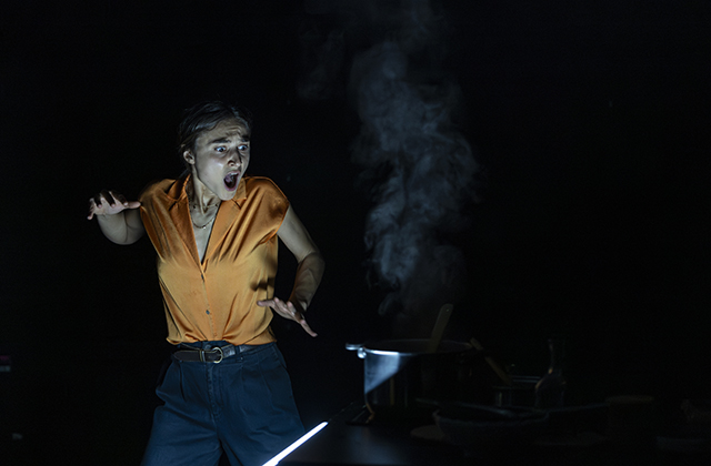 Isabella Nefar, in My English Persian Kitchen. The woman stands with a shocked look on her face, holding a pot that has a plume of steam coming off it. She is wearing a bright orange shirt which is contrasted against a very dark background.