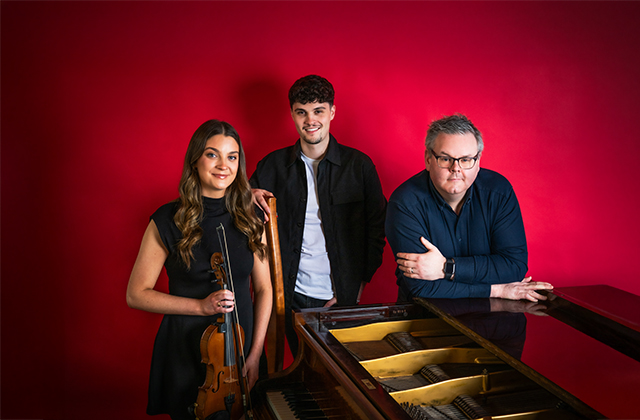 Séamus & Caoimhe Uí Fhlatharta, and Ryan Molloy all standing around a piano. Caoimhe is holding a violin