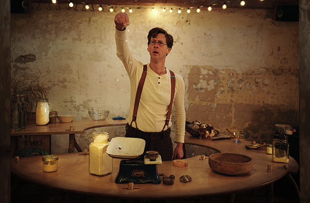 Manchán Magan at a weighing scales gesticulating as if explaining something. He wears suspenders and a white, longsleeve t-shirt.