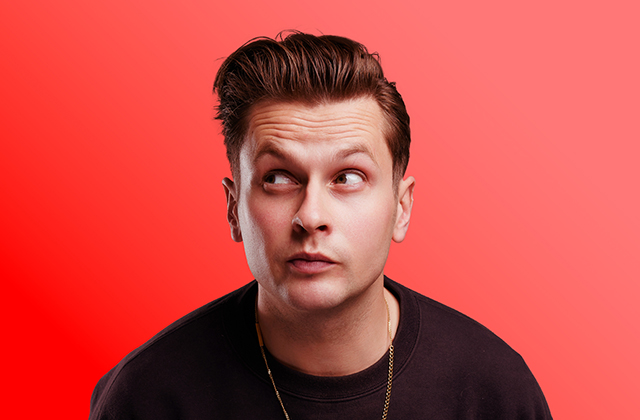 Headshot portrait of Mark Mehigan set against a vibrant solid red background. He is looking upward and off to the side with an expressive, quizzical look, styled with swept-back brown hair, wearing a dark sweater and a thin gold chain.