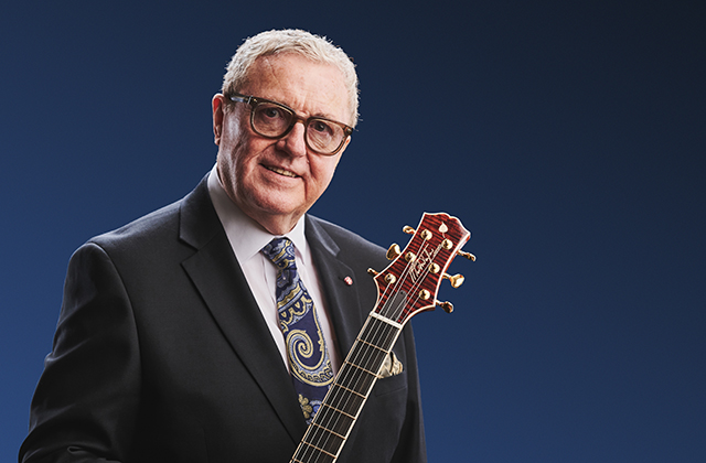 Martin Taylor, an older man in a dark suit and patterned tie, holding a guitar against a blue background.