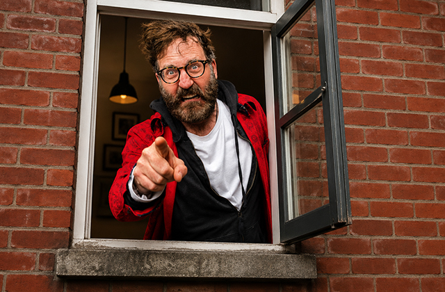 McSavage, a middle-aged man with wild, messy dark hair, glasses, and a full beard, leans out of an open ground-floor window set in a red-brick wall. He makes an exaggerated, intensely angry face while pointing directly forward at the camera. He wears a white t-shirt under a black hoodie and a bright red jacket.