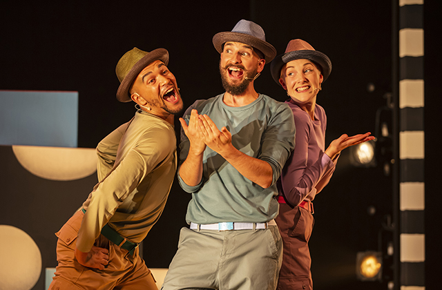 Human cast of the show appearing to be mid-song, dancing together, wearing hats and smiling while singing.