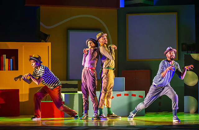 Two of the human cast stand back to back centre stage, they are flanked either side by other human cast members, each actor is making an animal like growling action with their hands.