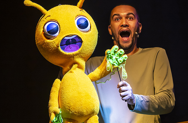 A large yellow alien like monster looks towards the camera with his human puppeteer, both of them look very surprised!
