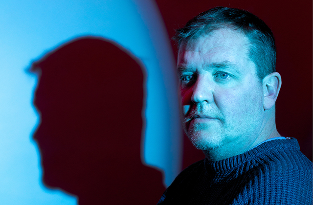 Garrett Lombard stands in a blue tinged spotlight, casting a striking shadow on the wall behind him
