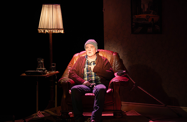 Garrett Lombard is pictured on the set of My Real Life by Eoin Colfer. He sits in a leather armchair, next to a table lamp. He is wearing a beanie hat, plaid shirt and cardigan. The background is dark and his face is lit up and he hold a thoughtful, worried expression on his face.
