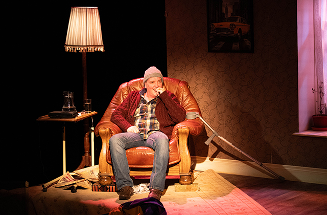 Garrett Lombard is pictured on the set of My Real Life by Eoin Colfer. He sits in a leather armchair, next to a table lamp. He is wearing a beanie hat, plaid shirt and cardigan. The background is dark and his face is lit up. he is smiling with his hand held to his cheek