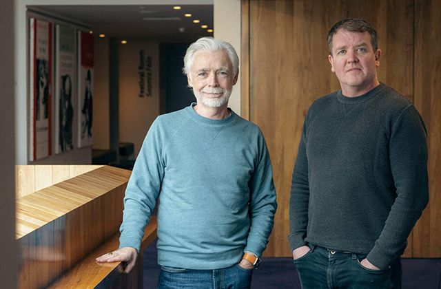 Eoin Colfer & Garrett Lombard are pictured together in the Foyer of a Theatre, they are standing towards the camera in jeans and jumpers and smiling.