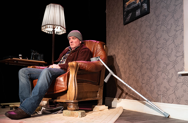Person sits in a leather armchair beside a floor lamp, with a crutch resting nearby on a detailed living-room theatre set.