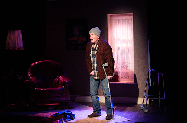 Person wearing a beanie stands on stage with a crutch, illuminated by theatrical lighting in a living-room setting.