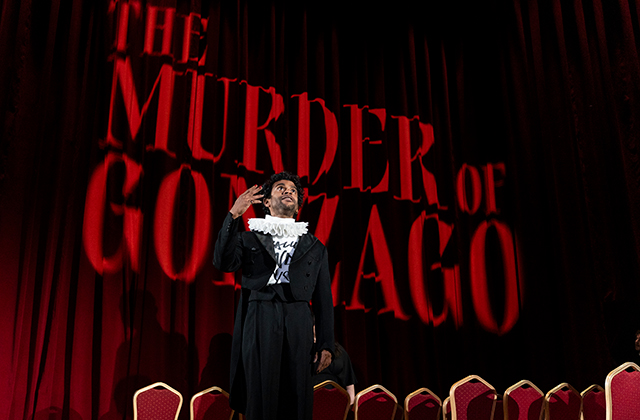 A stage scene with deep red curtains and the large projected words “The Murder of Gonzago” in dramatic red lettering. Hamlet (Hiran Abeysekerain) is in an Elizabethan-style black costume with a white ruffled collar standing center stage, raising a hand in a theatrical gesture, with a row of empty red chairs visible in the foreground.