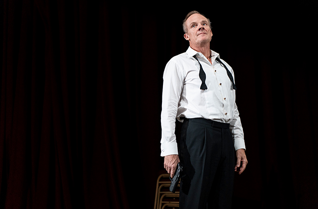 Alistair Petrie as Claudius. Claudius stands against a black background in a black tuxedo, his tie is undone, his suit jacket is off, he is holding a gun down by his side.