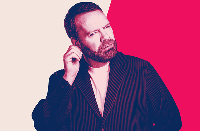 Neil Delamere with a puzzled expression scratches his ear, appearing to listen closely or think through a question.