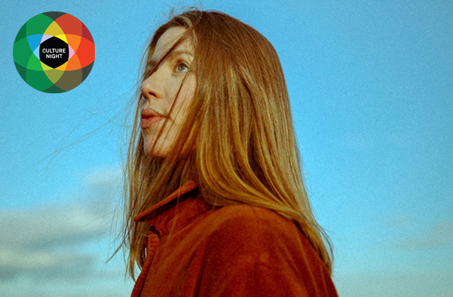 Niamh Regan stands with a blue sky behind her, wearing a red turtle neck, and her red hair windswept across her face