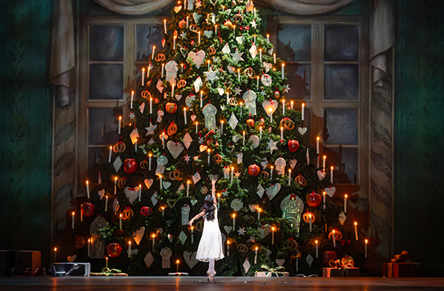 A stage production scene from The Nutcracker featuring a young ballet dancer seen from behind in a white dress, standing on pointe in front of a massive, towering Christmas tree. The tree is lavishly decorated with glowing candles, red baubles, star ornaments, and gingerbread-style decorations. Wrapped presents sit on the floor around the base, framed by large classic windows set into a dark green backdrop.