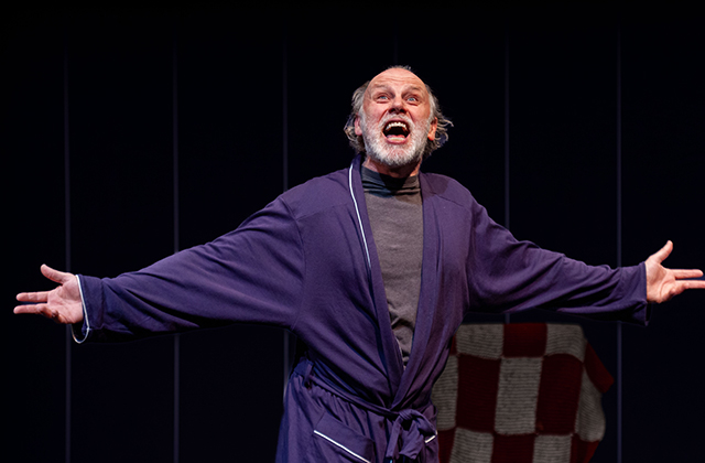 Don Wycherley in a blue robe, arms wide, with a wild expression on his face.