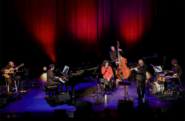 A live musical ensemble performing on a stage illuminated by warm red spotlighting against a dark background. The band includes a seated classical guitarist on the far left, a pianist at a grand piano, a vocalist in an orange top at center mic, an upright bassist behind her, a flutist, and a drummer on the far right.
