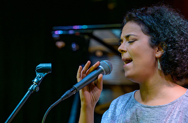 A close-up profile shot of a female singer with short curly dark hair and round glasses, holding a microphone to her mouth with eyes closed in focused performance. She wears dangling earrings and a light-colored top, with the dark background softly blurred.