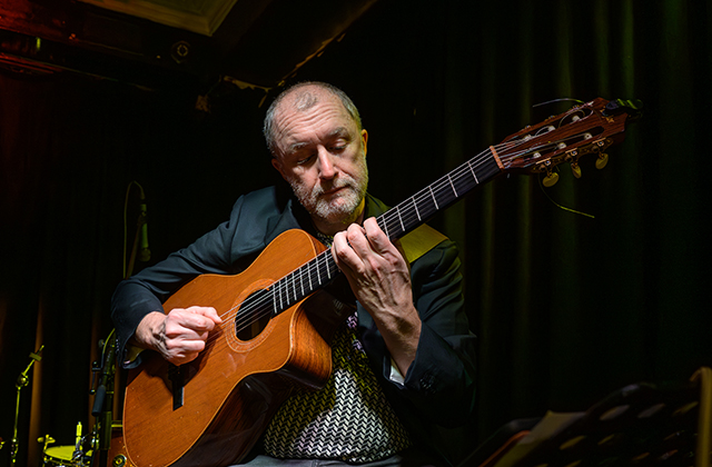 A close-up shot of a male musician playing a nylon-string acoustic guitar on a dimly lit stage. He has short grey hair and a beard, wearing a dark blazer over a patterned shirt, holding the guitar neck with focused concentration under warm, subtle stage lighting.