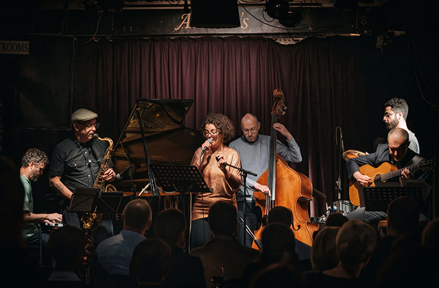 A full live ensemble performing on an intimate stage with deep red velvet curtains in the background. The group features a pianist, a saxophonist wearing a flat cap, a lead female vocalist at center, a double bass player, an acoustic guitarist, and a percussionist, with the dark silhouettes of the audience visible in the foreground.