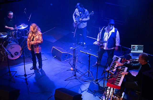 Live band performing on a dimly lit stage under blue lighting. A female vocalist stands centre with a microphone, surrounded by musicians: a drummer at the back, a guitarist and saxophonist to the right, and a keyboardist in the foreground. Instruments and monitors are arranged across the stage, creating an intimate concert atmosphere.