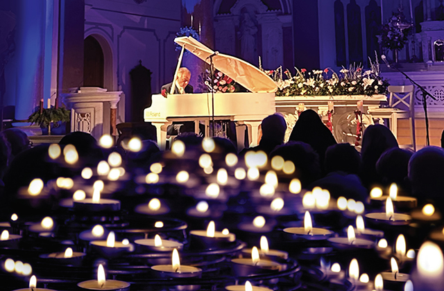 Phil Coulter playing a white grand piano in front of a silhouetted audience, and a sea of candles