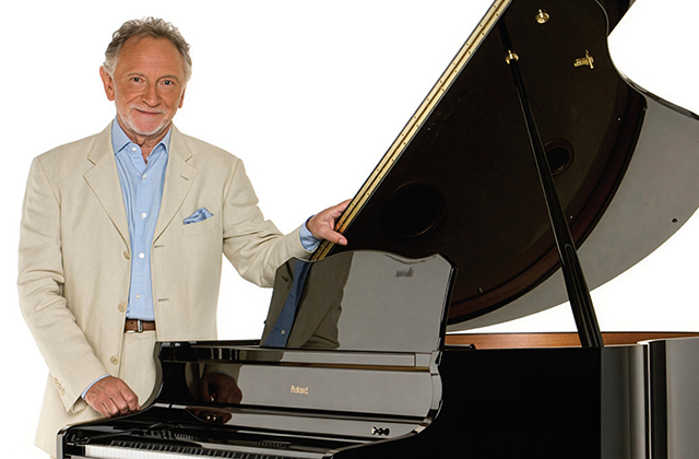 Phil Coulter in a cream blazer and light blue shirt stands beside a glossy black grand piano with the lid open, smiling at the camera.